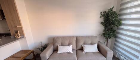 A living room with a beige sofa, a wooden bench, a potted plant, and a window with blinds.