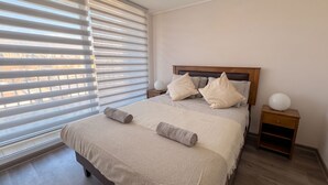 Una cama bien tendida con almohadas y una toalla enrollada, una mesita de noche con una lámpara, y una ventana con persianas.
