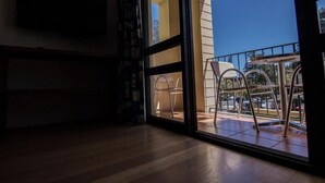 Una habitación con suelo de madera, televisor montado en la pared y balcón con sillas y mesa de metal.