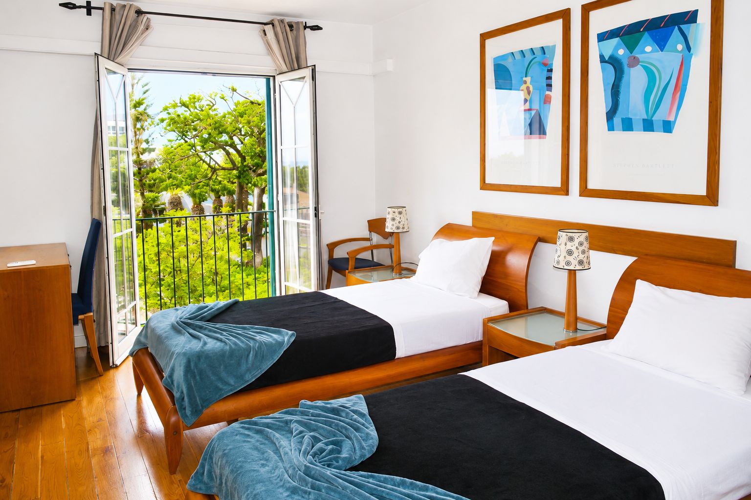 A hotel room with two beds, a balcony with a view of greenery, and two framed artworks on the wall.