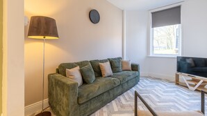A green sofa with cushions, a floor lamp, a clock, a TV on a stand, and a window with blinds. - Anika in South Shields (South Shields)