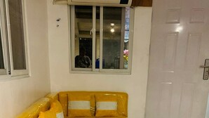 A yellow sectional sofa with white accent pillows, a window with blinds, a white door, and a tiled floor.