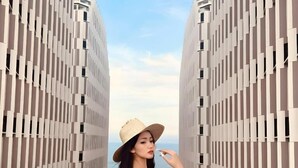 Una mujer con un vestido estampado y sombrero de paja junto a una piscina, con edificios modernos al fondo.