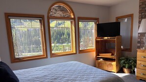 A bedroom with a bed, a TV, a dresser, and three windows with a view of a forest and mountains.