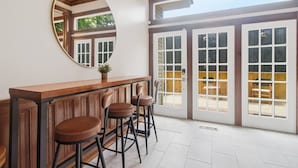 Un espace bar avec un comptoir en bois et trois tabourets, de grandes fenêtres à carreaux et un grand miroir rond accroché au mur.