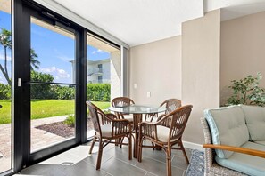 A modern living room with a glass table and wicker chairs, a light-colored sofa, and a view of a green garden and buildings. - Sanibel Sunrise 3 bed 3 bath Beachfront Condo (Sanibel)