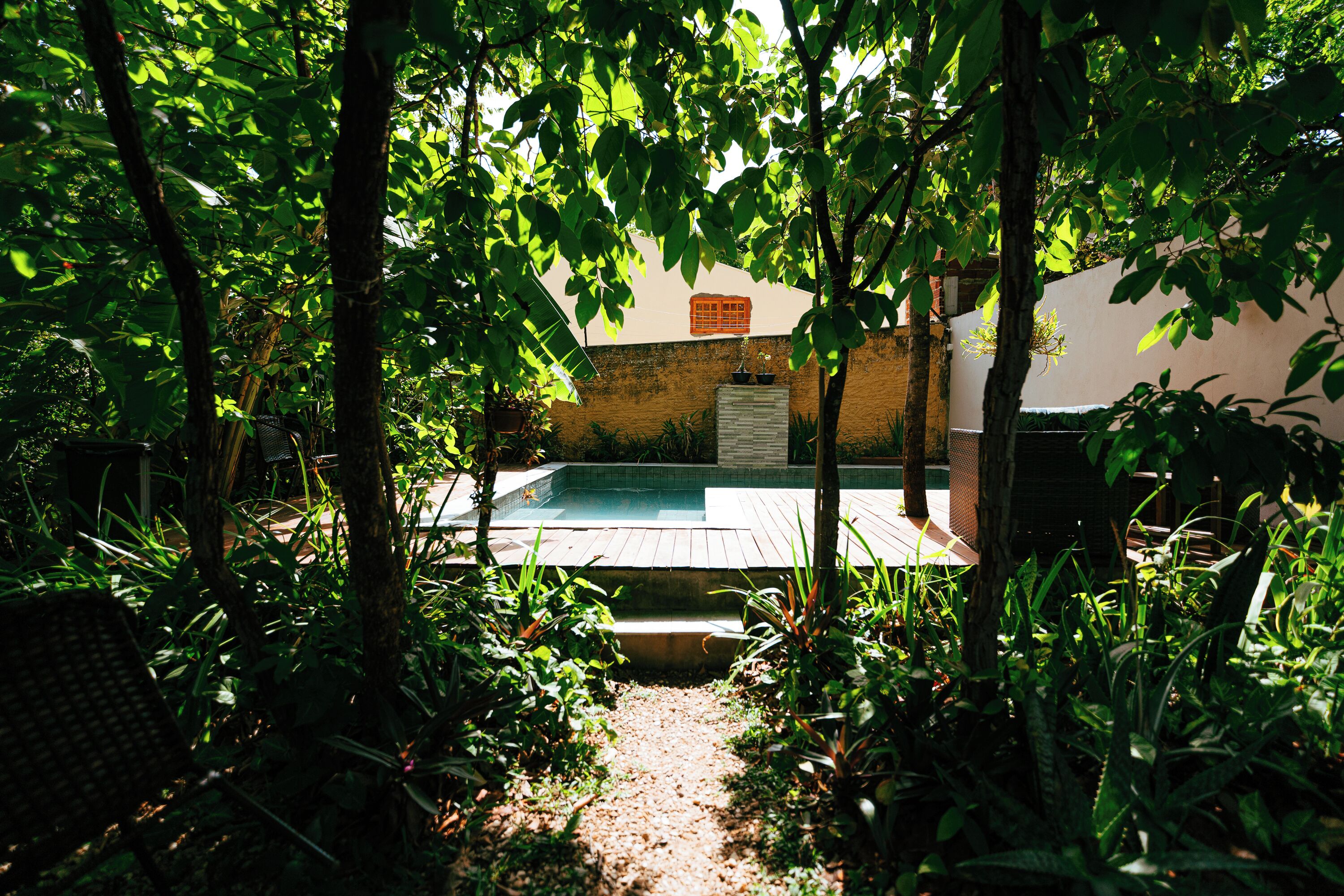 Un área de piscina rodeada de vegetación frondosa, una terraza de madera y un edificio con pared color beige y una puerta.