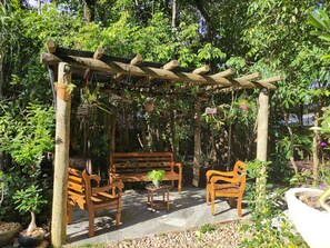 A wooden pergola with benches and hanging plants in a lush garden setting. - Pousada Templários Ecovilla  (Pirenópolis)