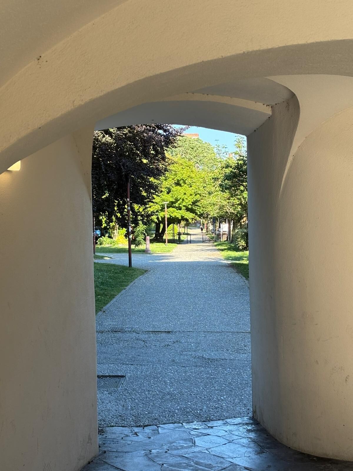 An arched corridor with a paved path leading to a park area with trees and a clear sky.