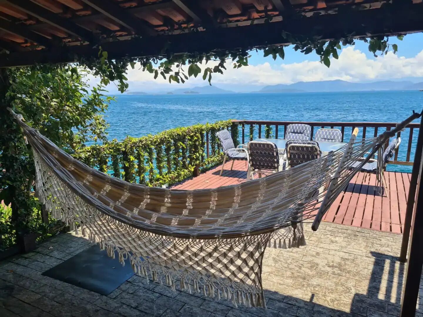 Uma rede sob uma pérgola, uma mesa com cadeiras e vista para o mar.