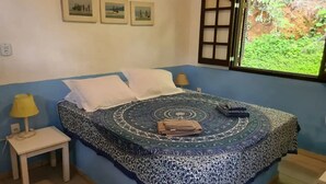 A bedroom with a bed, a nightstand, a window with a view of greenery, and three framed pictures on the wall.