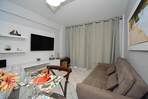 Una sala de estar con un sofá, un televisor montado en la pared, una mesa de comedor con sillas y un ventanal con cortinas.