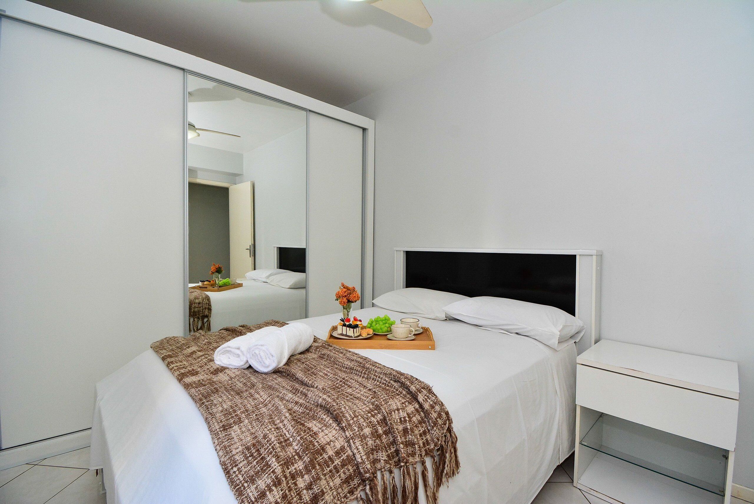 Una habitación de hotel moderna con una cama grande, una mesita de noche, un espejo y una bandeja con pastel y fruta.