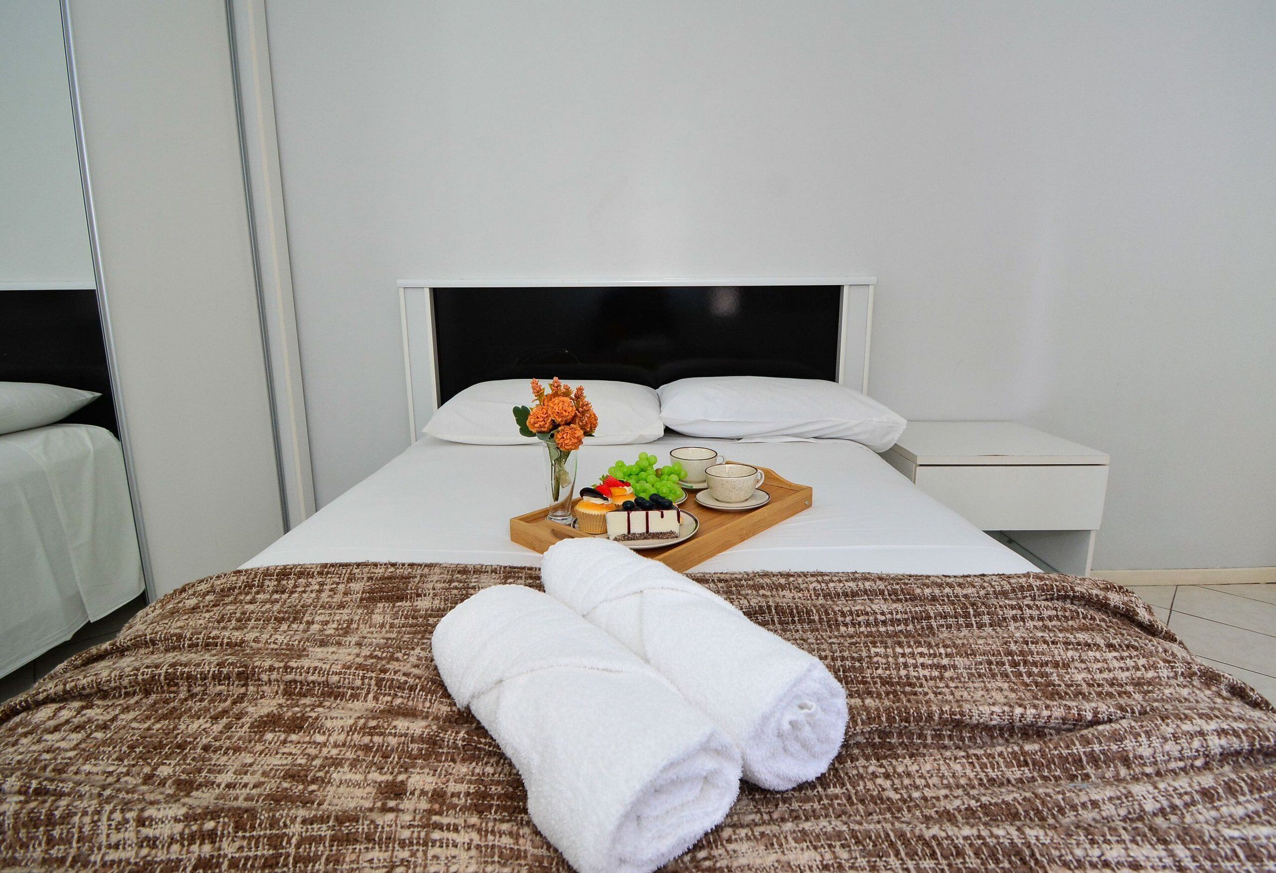 Una cama bien tendida con una colcha marrón y blanca estampada, dos toallas blancas enrolladas, una bandeja con frutero, un pastel pequeño y un jarrón de flores.