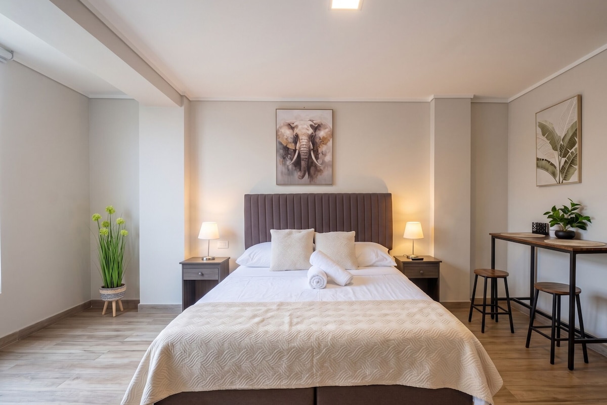 A hotel room with a bed, bedside tables, a desk with two chairs, a potted plant, and artwork on the walls.