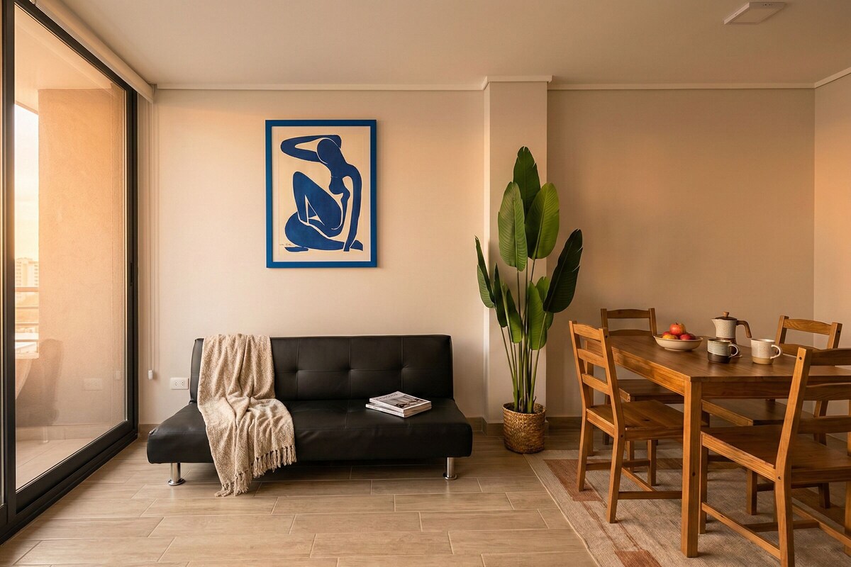A modern living room with a black sofa, a dining table with wooden chairs, a potted plant, and a framed abstract artwork on the wall.