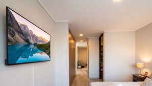A hotel room with a large flat-screen TV mounted on the wall, a neatly made bed with white linens, a bedside table with a lamp, and a view of a mountain landscape through the window. - Barrio París Londres - Domus Travels (Santiago)