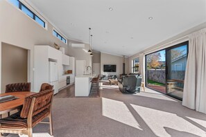 A spacious living area with a dining table, sofa, and TV, connected to a kitchen and a sliding glass door to the outdoors. - Riverstone Retreat - Turangi Holiday Home (Turangi)
