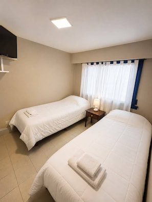 Habitación de hotel con dos camas, un televisor de pantalla plana montado en la pared, una mesita de noche con lámpara y una ventana con cortinas.