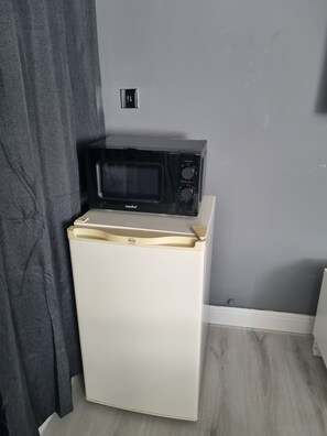 Een compacte kitchenette met een magnetron bovenop een witte koelkast, een klein spoelgedeelte en een grijze gordijn.