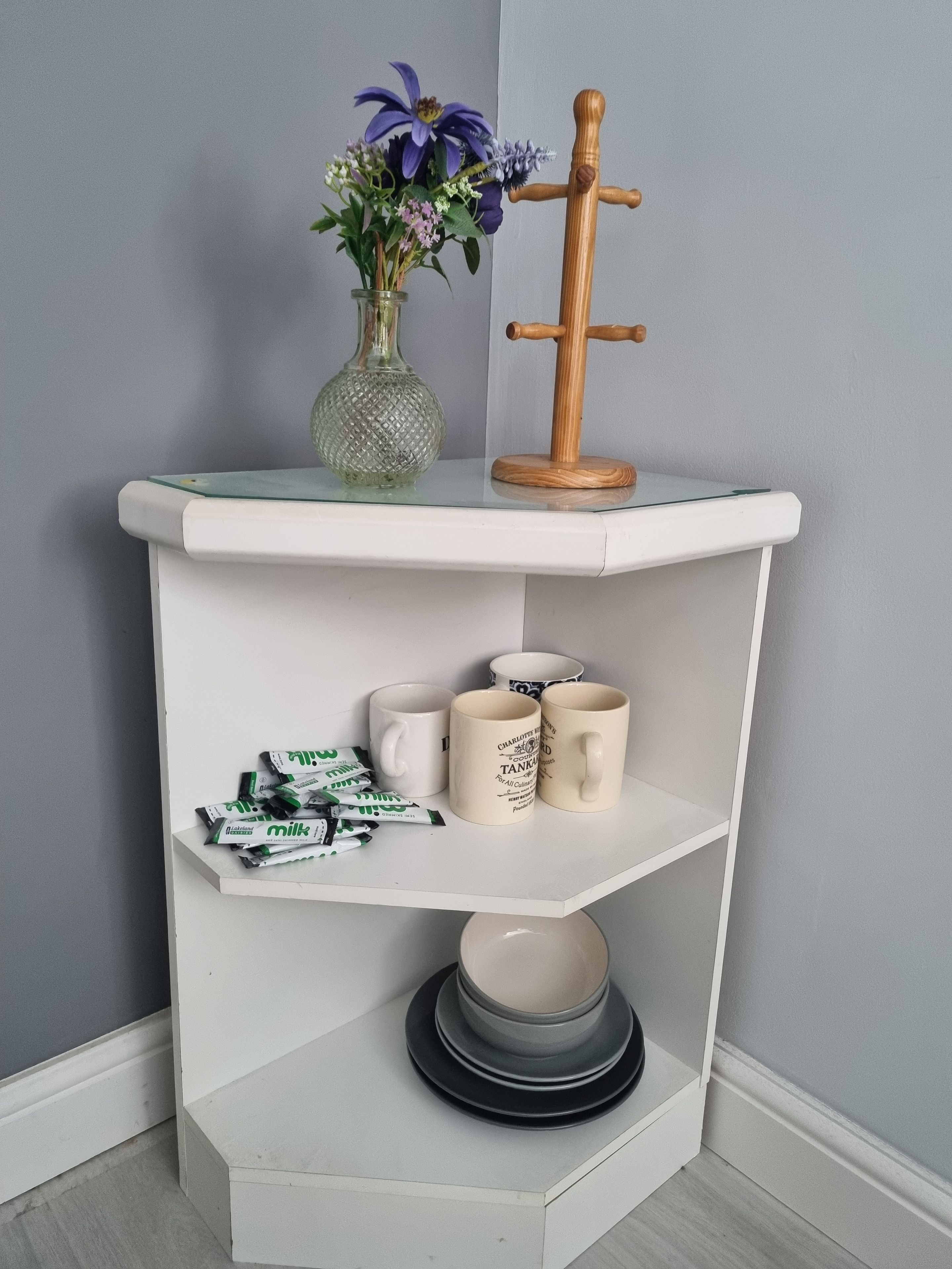 A corner shelf unit with a vase of flowers, a wooden decorative item, and a mug with text.