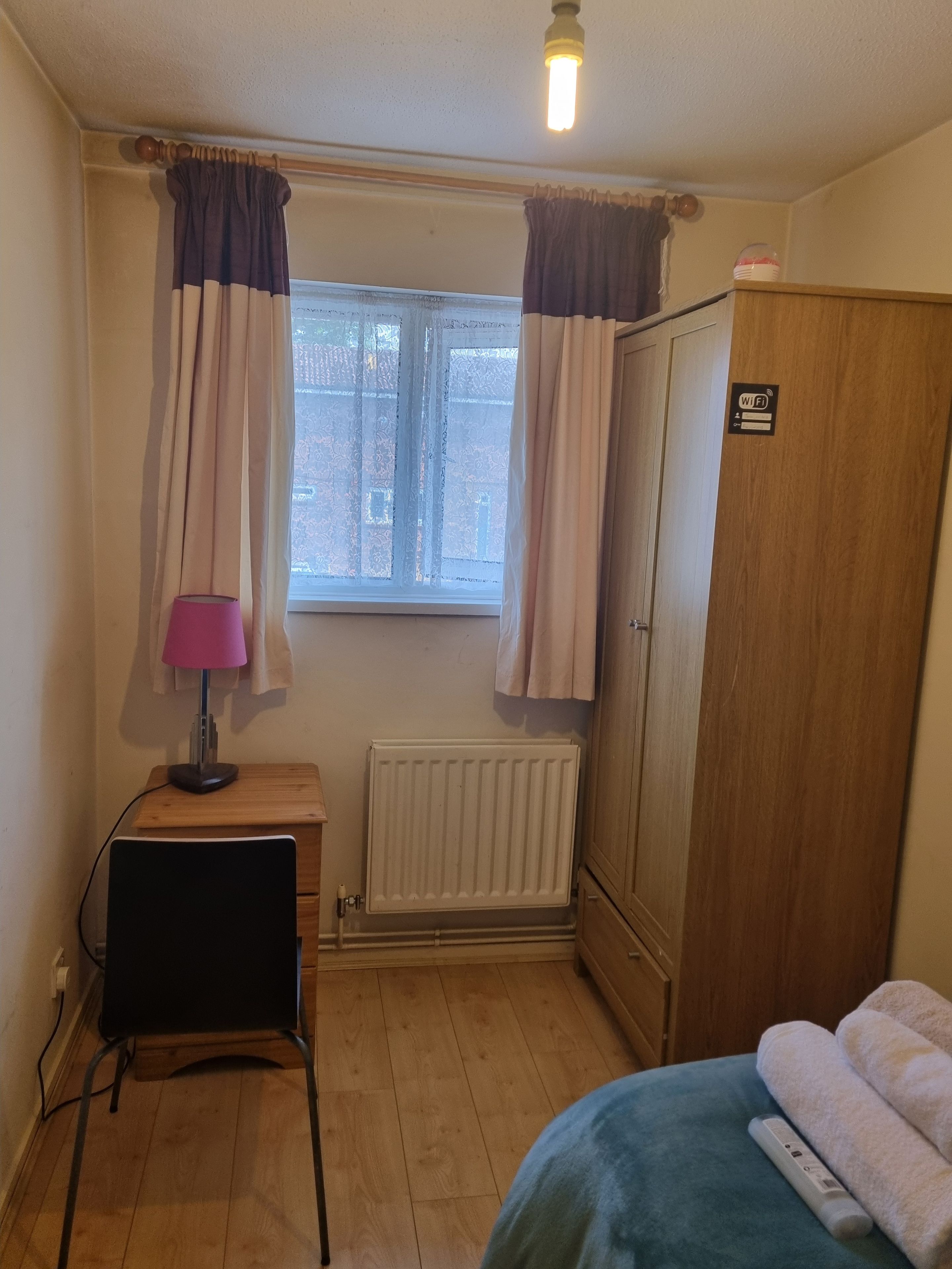 A room with a radiator, a desk with a lamp, a wardrobe, and a bed with a blanket.