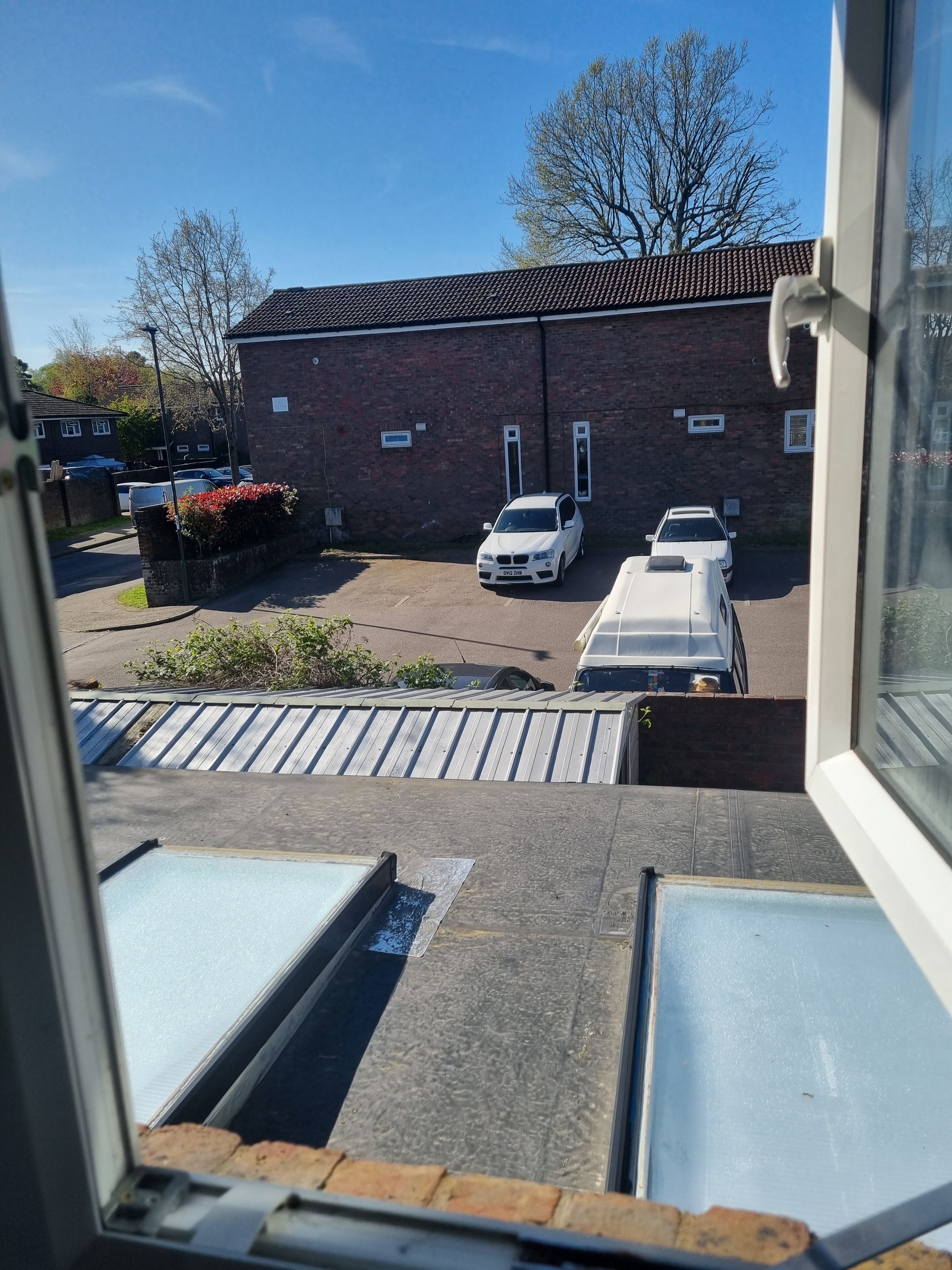 Une vue depuis une fenêtre donnant sur un parking où se trouvent deux voitures blanches, un bâtiment en briques et un ciel bleu dégagé.