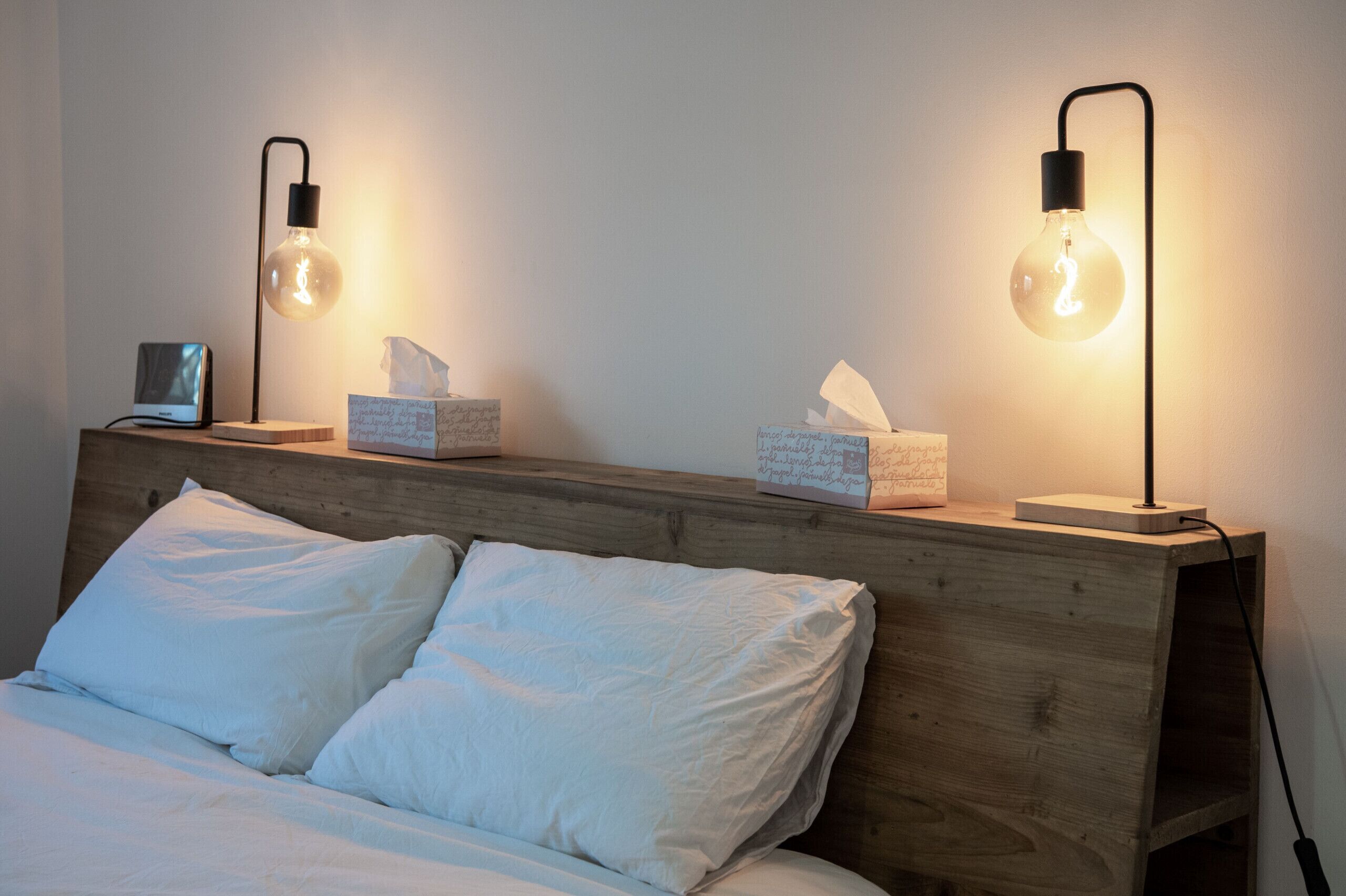 Een slaapkamer met een houten hoofdbord, twee bedlampjes, een nachtkastje met zakdoos en een klein elektronisch apparaat.