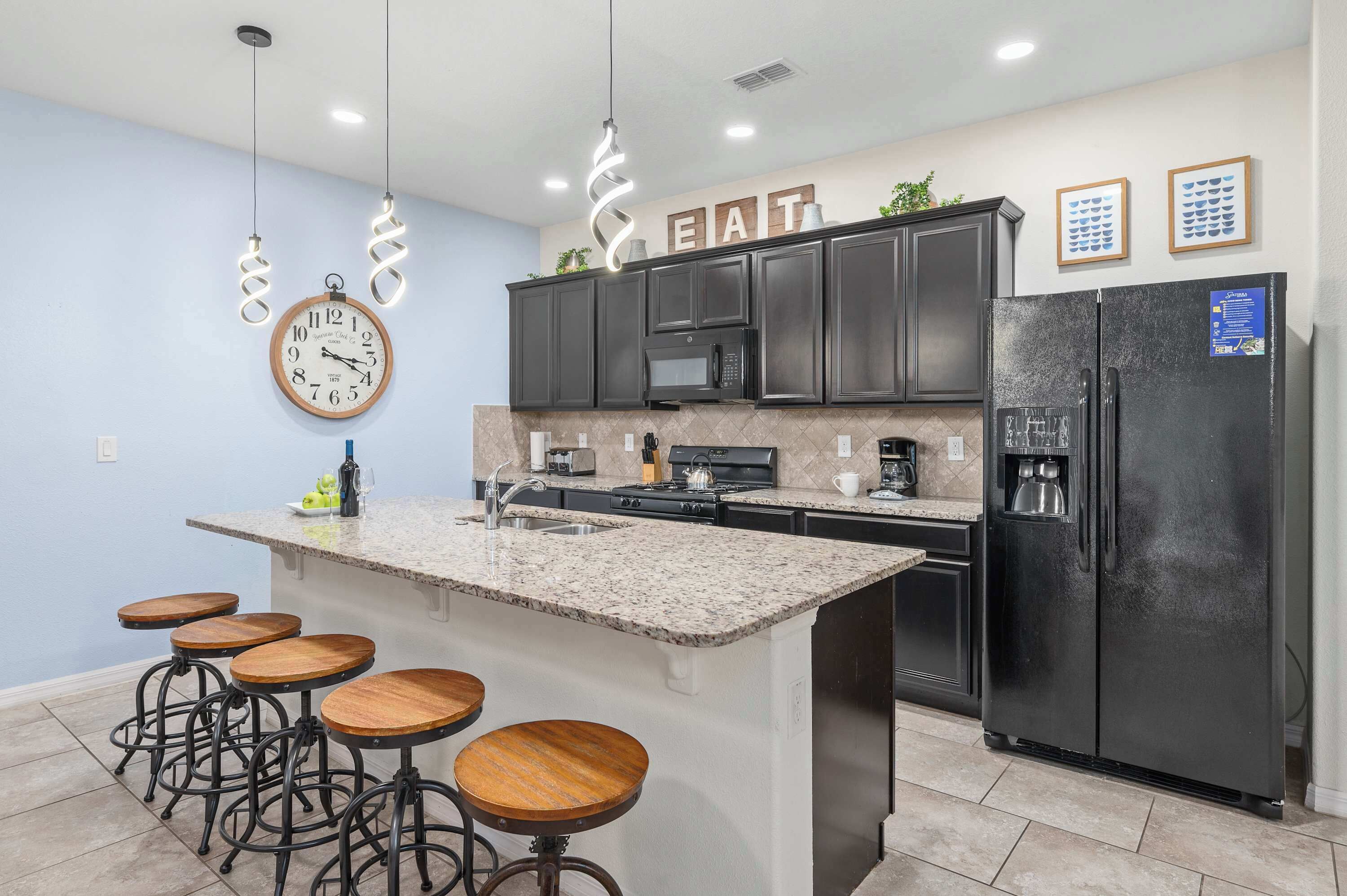 A modern kitchen with dark cabinets, a central island with stools, a refrigerator, and a clock on the wall.