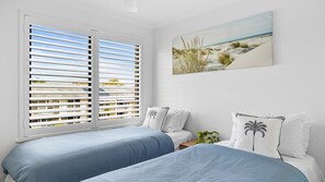 Ein Schlafzimmer mit zwei Betten, weißen Wänden, einem Gemälde mit Strandmotiv und einem Fenster mit Jalousien.