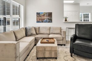 Una sala de estar con un sofá modular, un sillón de cuero negro, una mesa de centro de madera y un cuadro colgado en la pared que muestra una cascada.