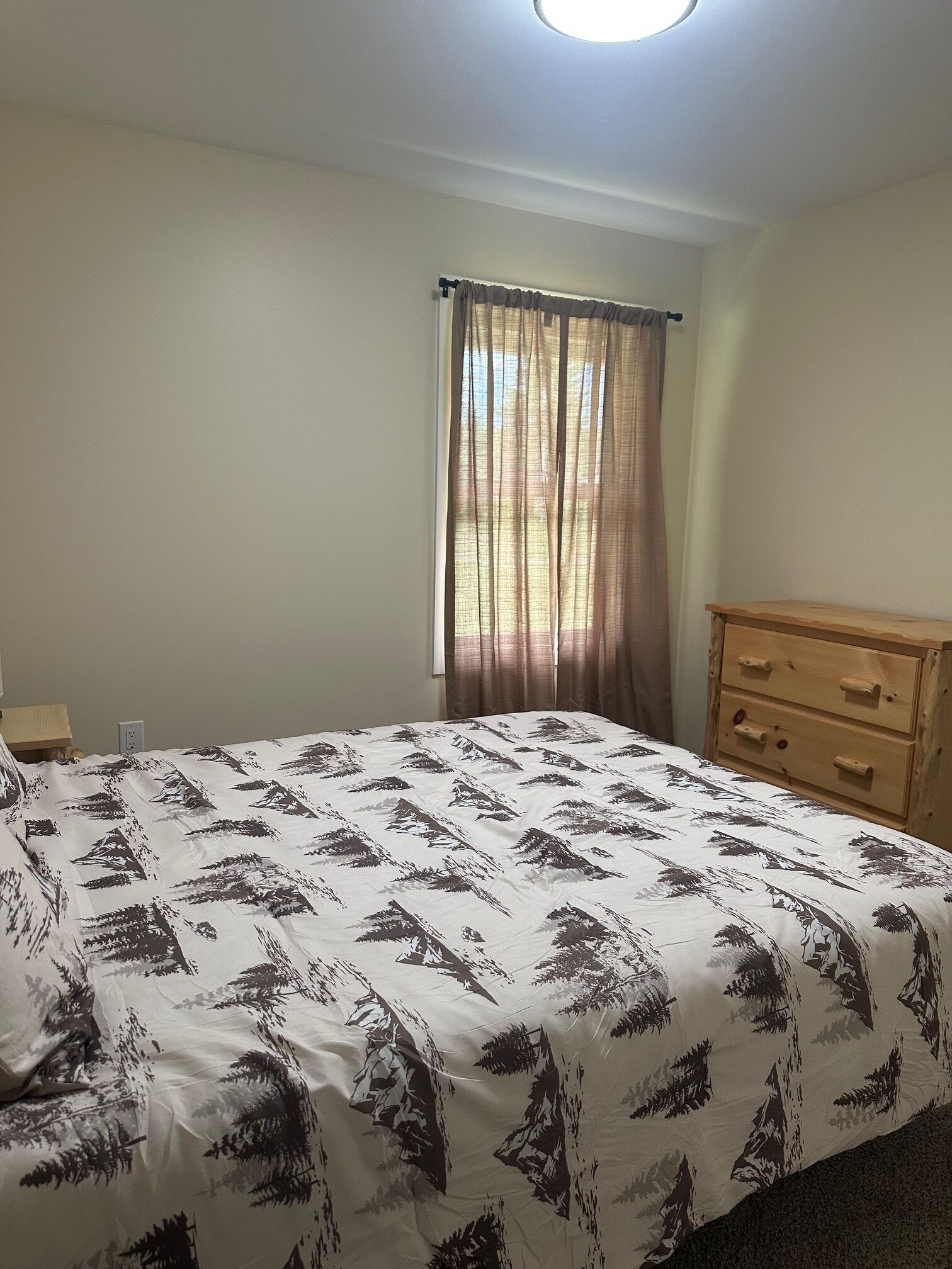 A bedroom with a bed, a wooden dresser, a window with curtains, and a wall socket.