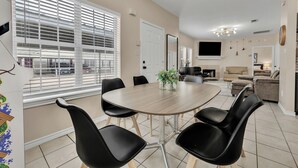A dining area with a round table and black chairs, a TV in the background, and a large window with blinds. - TikTok: Walk to Texas A&M Campus, Sleep 8, 3BR/3BA (College Station)