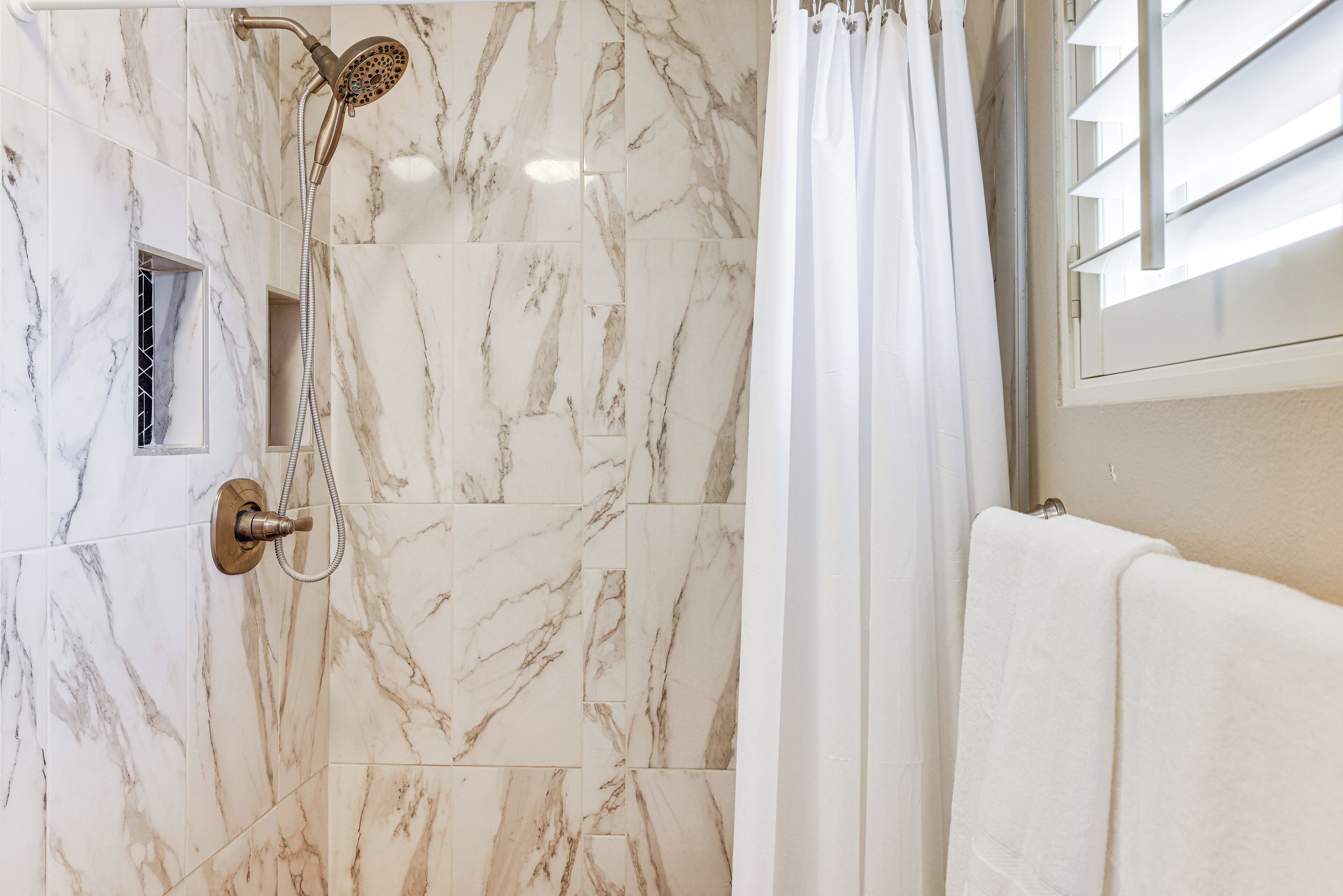 Une salle de bain carrelée de marbre, avec une douche, un porte-serviettes et une fenêtre équipée de stores.