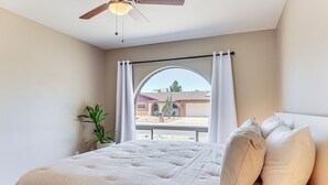 Ein Schlafzimmer mit Bett, Deckenventilator, Fenster mit Vorhängen und Blick ins Freie.