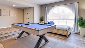 Ein Zimmer mit Billardtisch, einem Bett, einem großen Fenster mit Vorhängen und einer Zimmerpflanze.