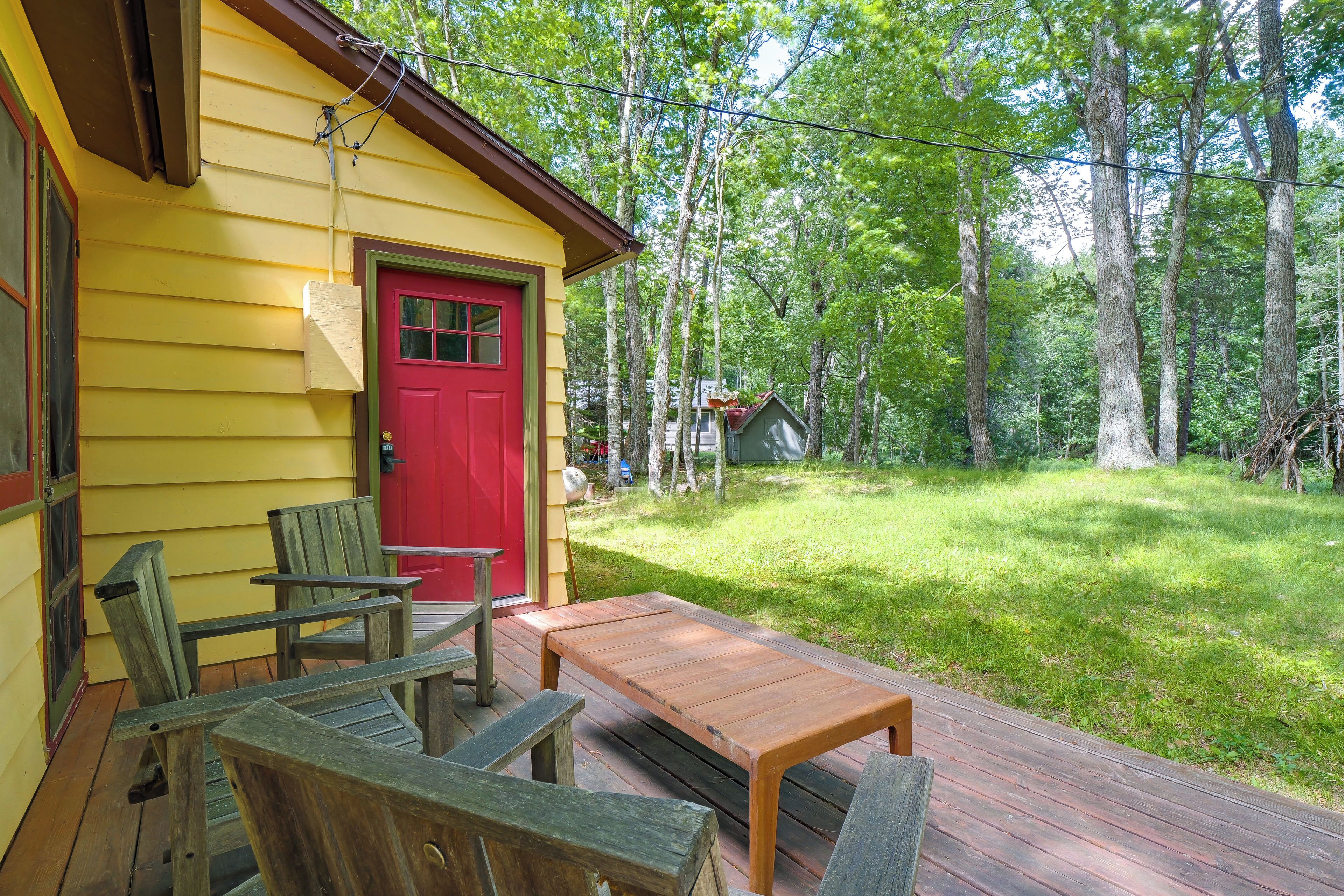 A yellow wooden house with a red door, a wooden deck, and a grassy yard surrounded by trees.