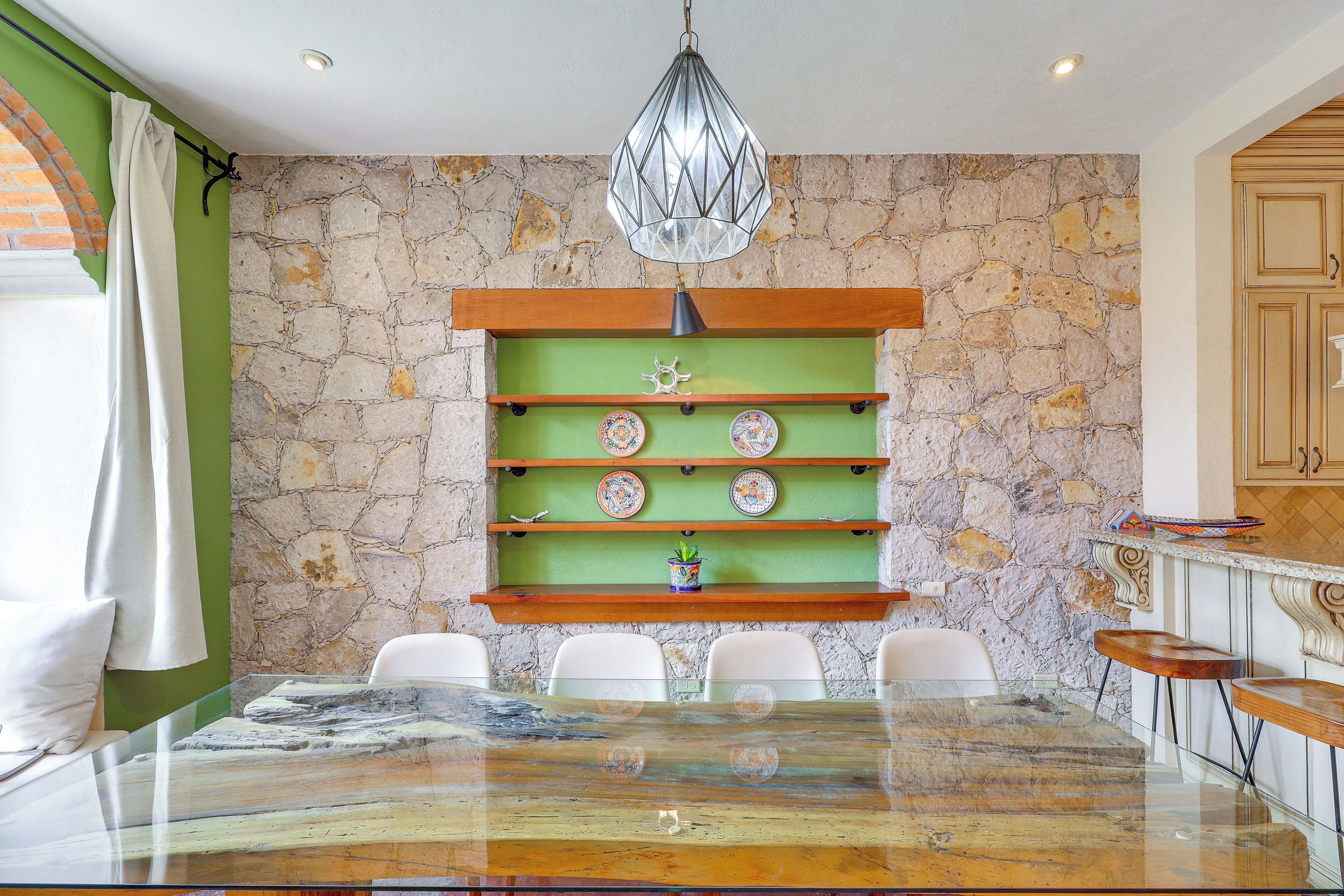 A dining area with a stone wall, a green shelf with decorative plates, a glass table, and white chairs.