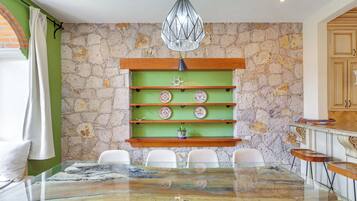 A dining area with a stone wall, a green shelf with decorative plates, a glass table, and white chairs.
