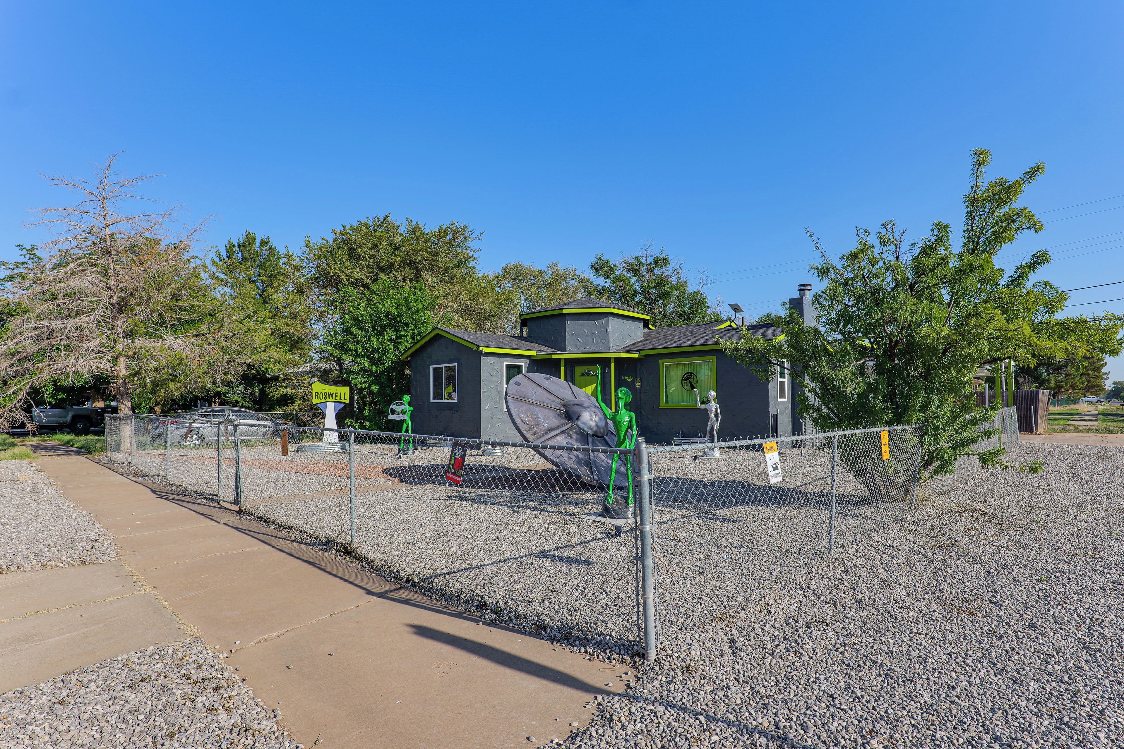 A small, modern house with a green roof, surrounded by a gravel area and a metal fence.