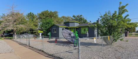 A small, modern house with a green roof, surrounded by a gravel area and a metal fence.