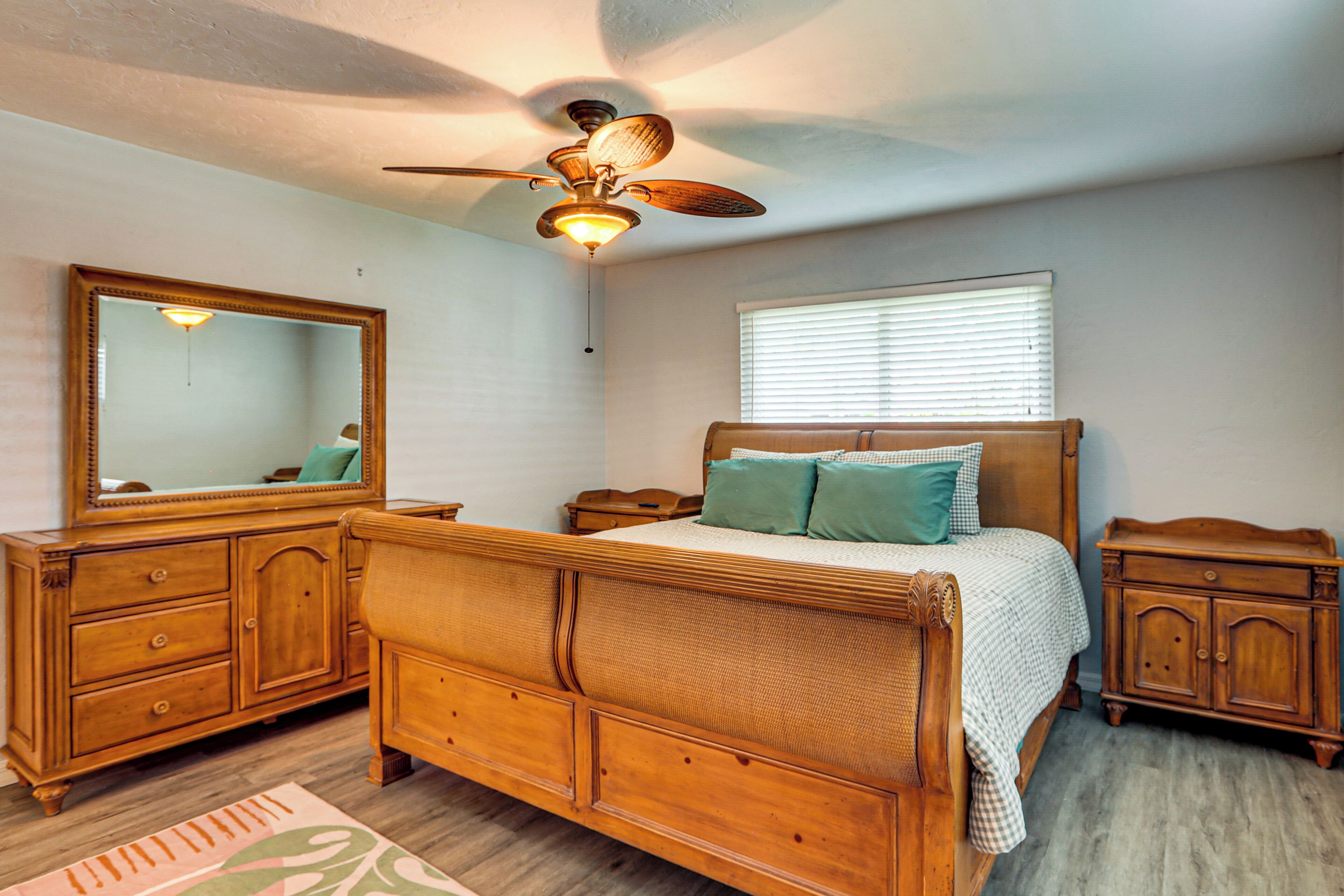 Une chambre à coucher avec un lit en bois, une table de chevet, une commode, un ventilateur de plafond, un miroir et une fenêtre avec des stores.