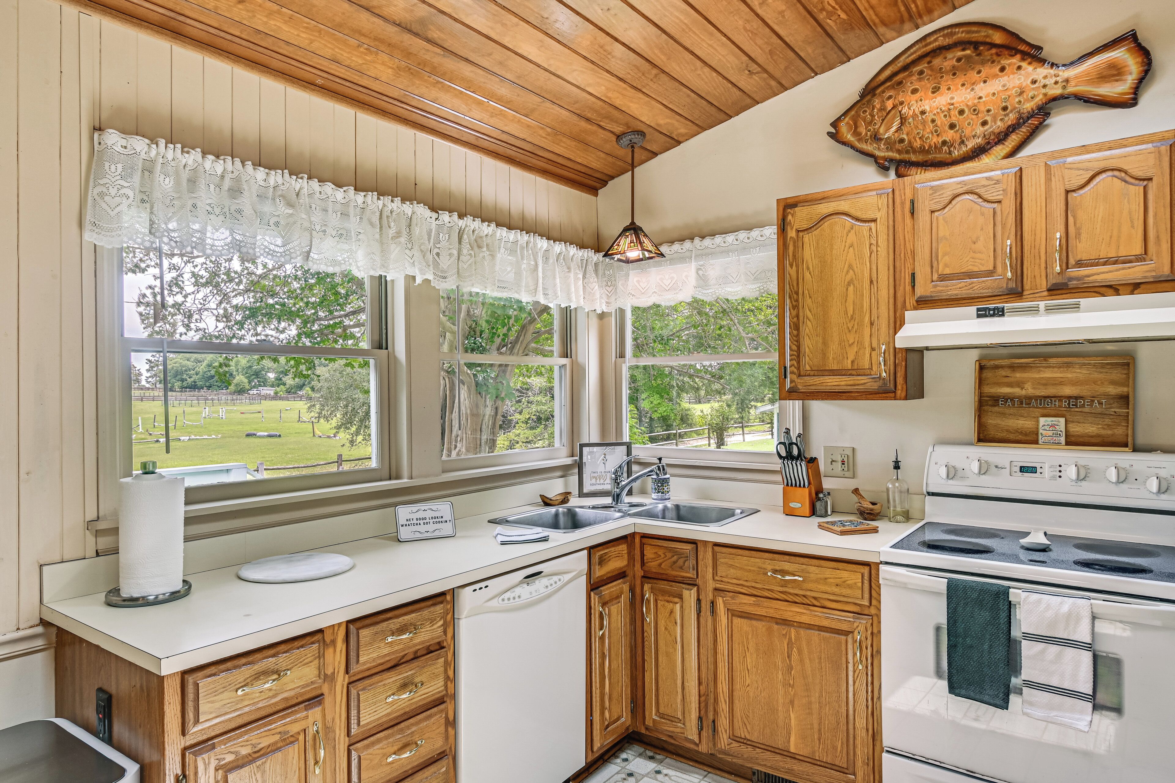 A kitchen with wooden cabinets, a white countertop, a stove, a sink, and a window with a view of a green landscape.