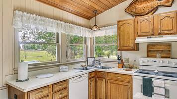 A kitchen with wooden cabinets, a white countertop, a stove, a sink, and a window with a view of a green landscape.