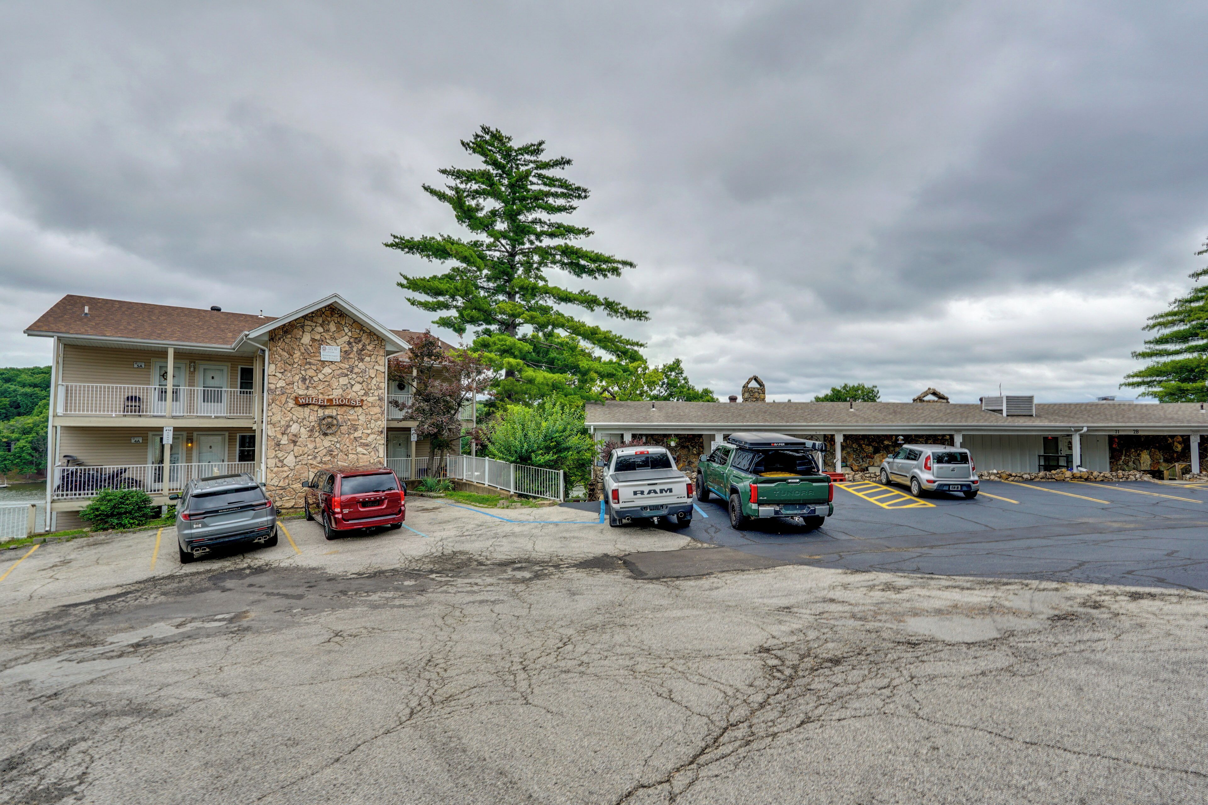 A motel with a stone facade, a parking lot with several vehicles, and a large tree in front.