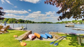 A lakeside setting with kayaks and paddleboards on the grass, a dock, and a view of the water and surrounding greenery.