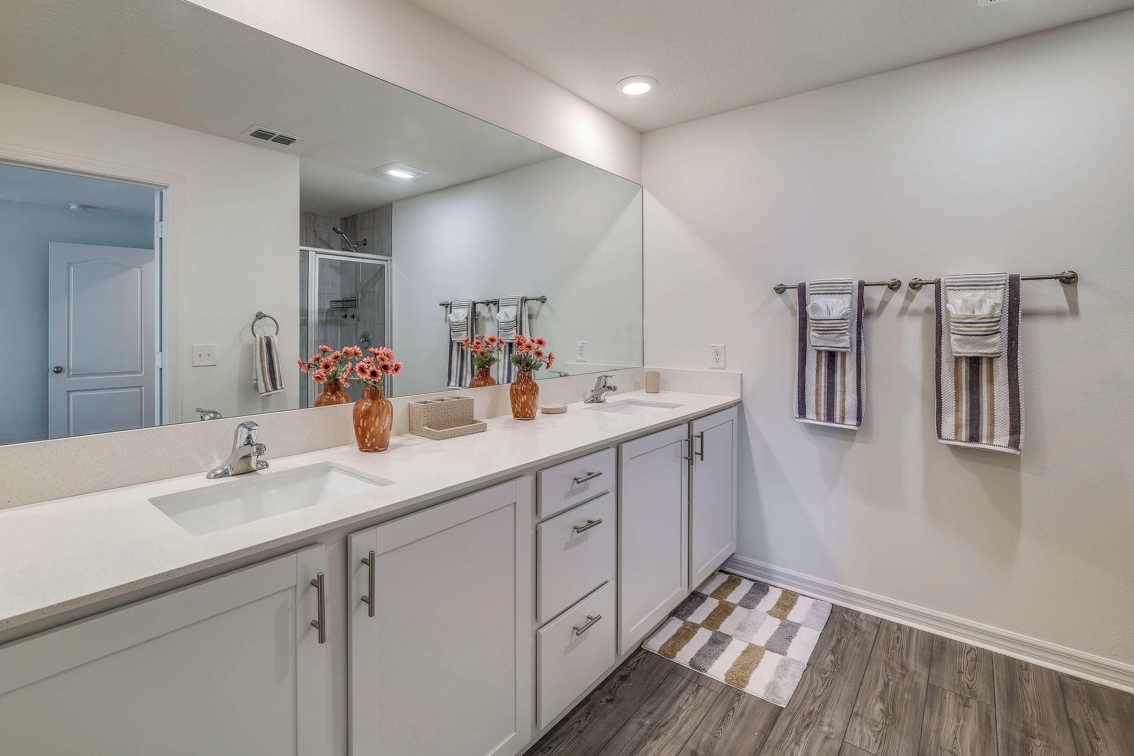 A modern bathroom with a large mirror, two sinks, a shower, and a patterned rug.