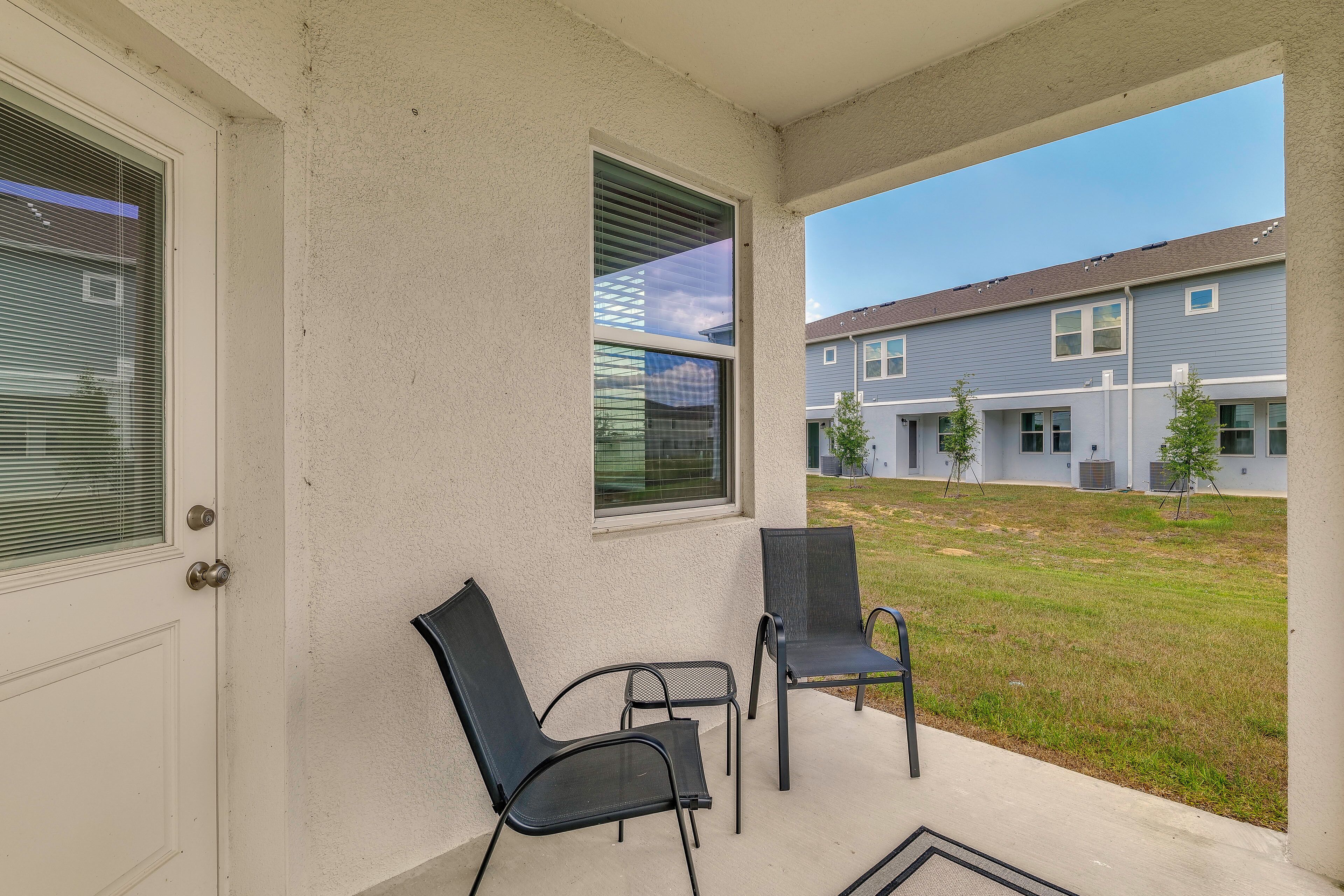 A covered porch with two chairs, a door, and a window overlooking a building complex.