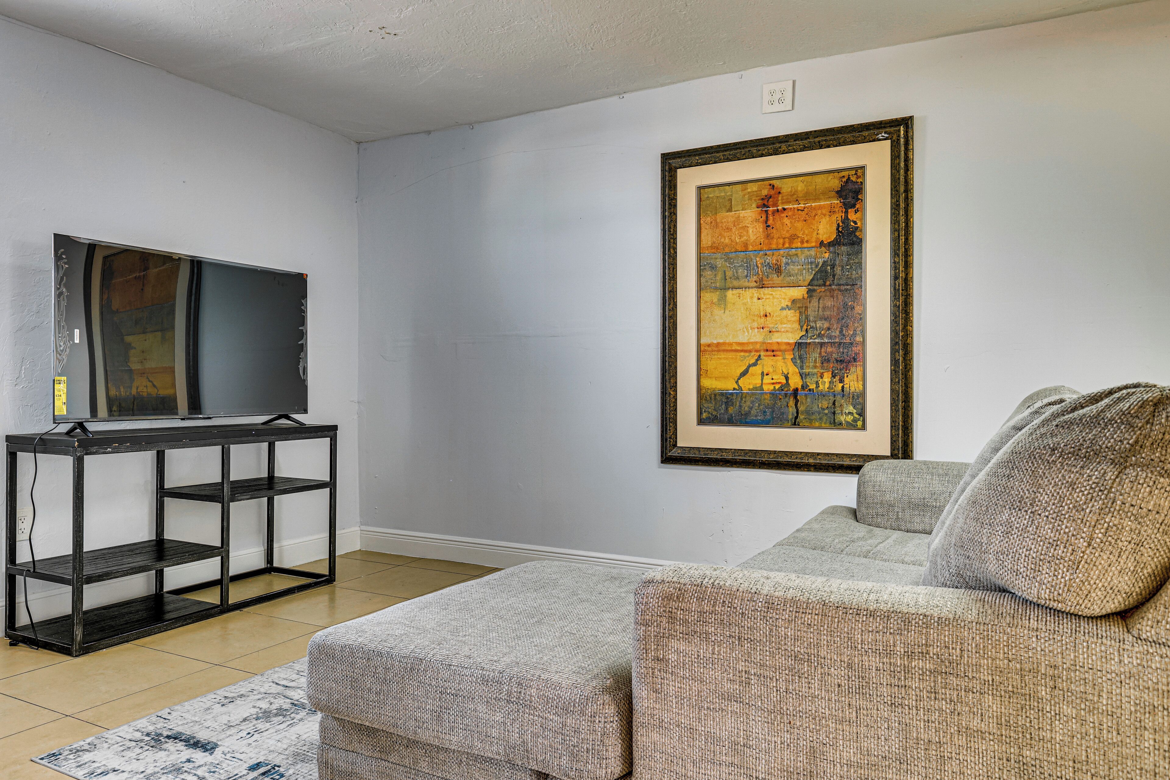 A living room with a sofa, a TV on a black metal stand, a framed painting on the wall, and a patterned rug.