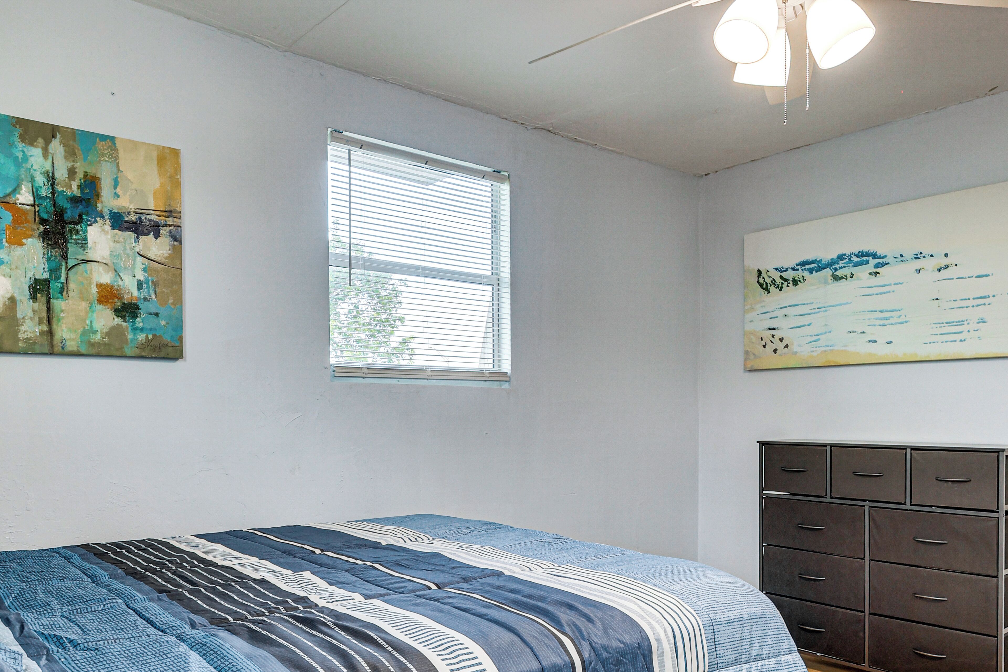 A bedroom with a bed, a window with blinds, a dresser, and two wall-mounted artworks.